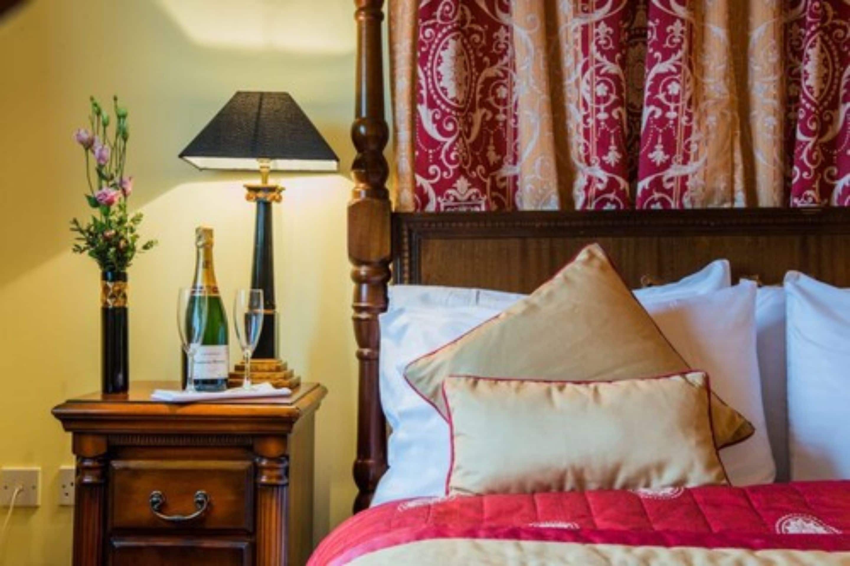 Une chambre d’hôtel avec une tête de lit en bois, une table de chevet avec une lampe, une bouteille de champagne et un vase rempli de fleurs.
