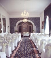 Un salón para eventos formales con filas de sillas blancas, una araña de cristal y una alfombra estampada.