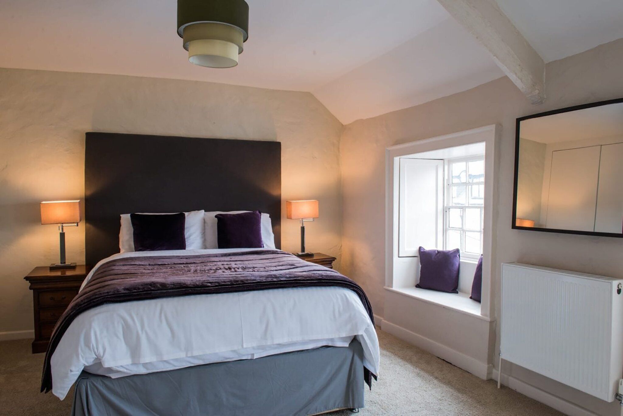 A bedroom with a large bed, two bedside lamps, a window with a view, and a radiator.