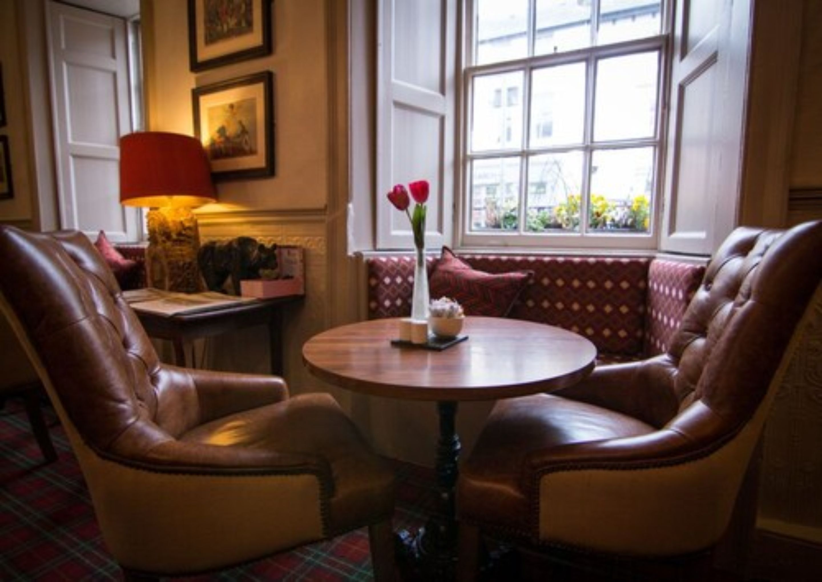 A cozy seating area with leather chairs and a round wooden table, featuring a vase with flowers and a red lamp.