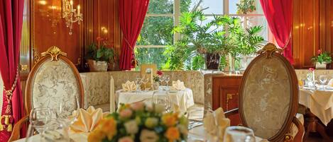 A dining area with tables set for a meal, a chandelier, and a view of greenery outside.
