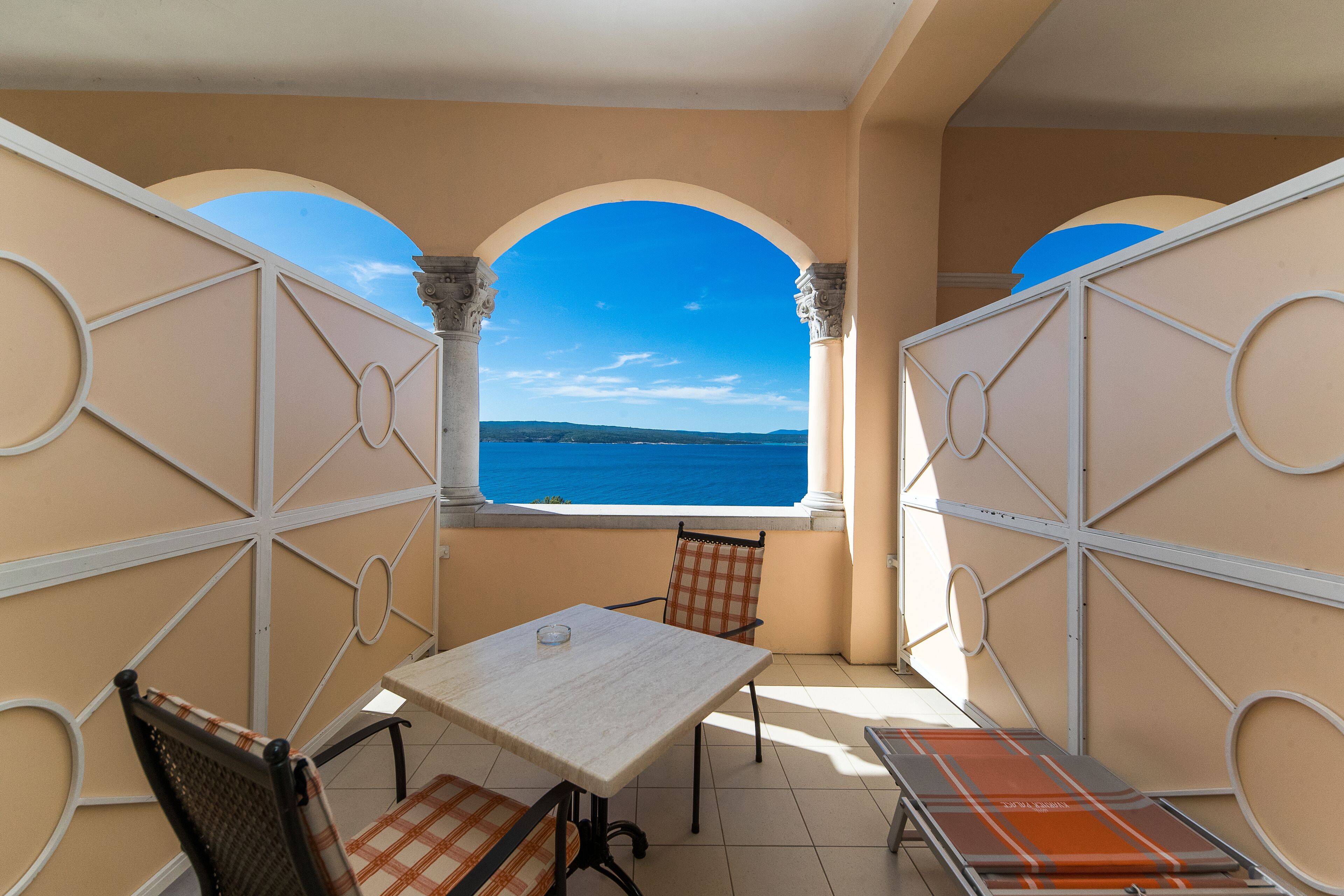A balcony with a table and chairs, a view of the sea, and a decorative wall with geometric patterns.
