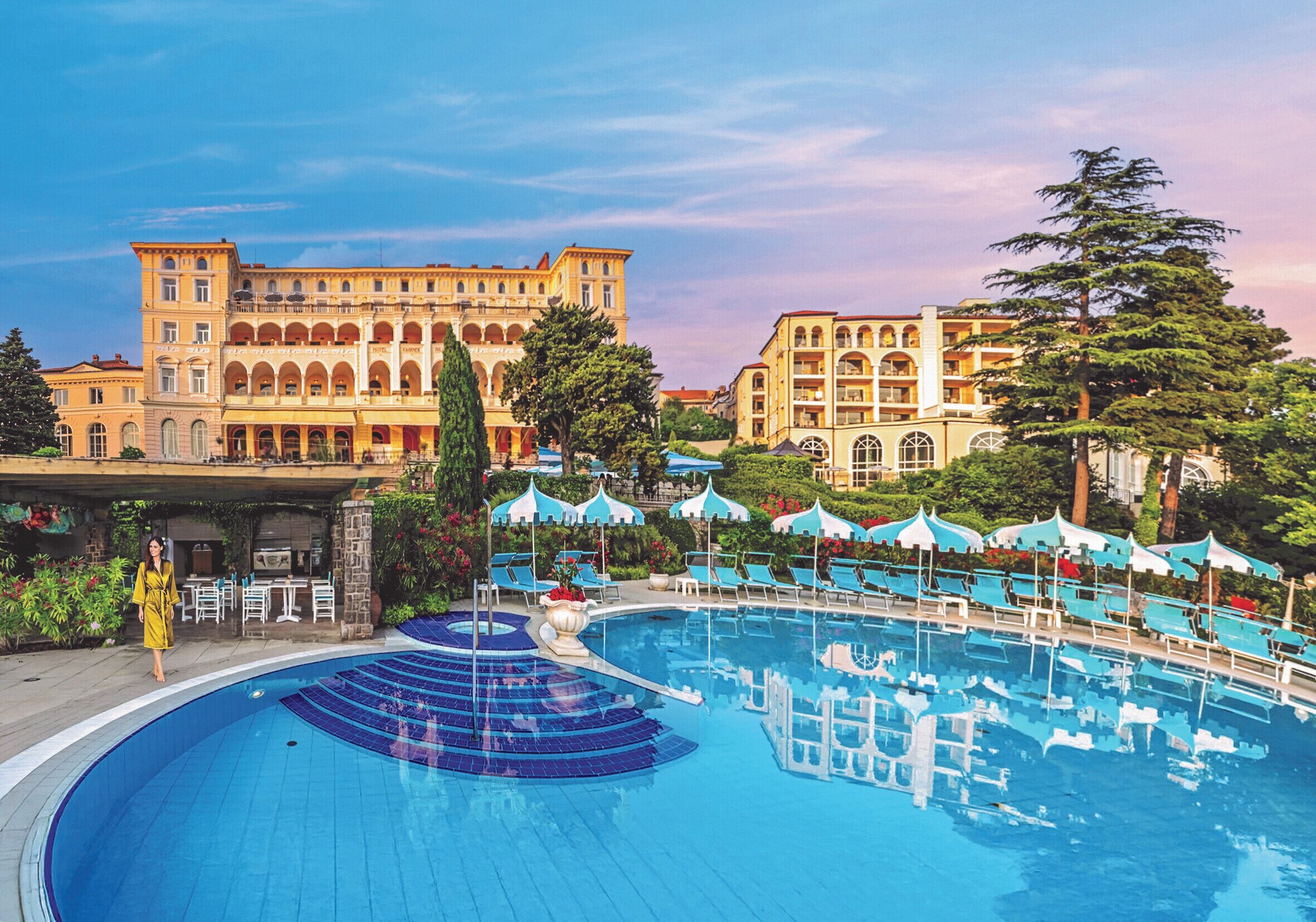 A large outdoor swimming pool with blue water, surrounded by lounge chairs and umbrellas, and a grand building with multiple floors and balconies in the background.