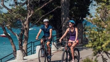 Two people riding bicycles on a paved path with trees and a body of water in the background.