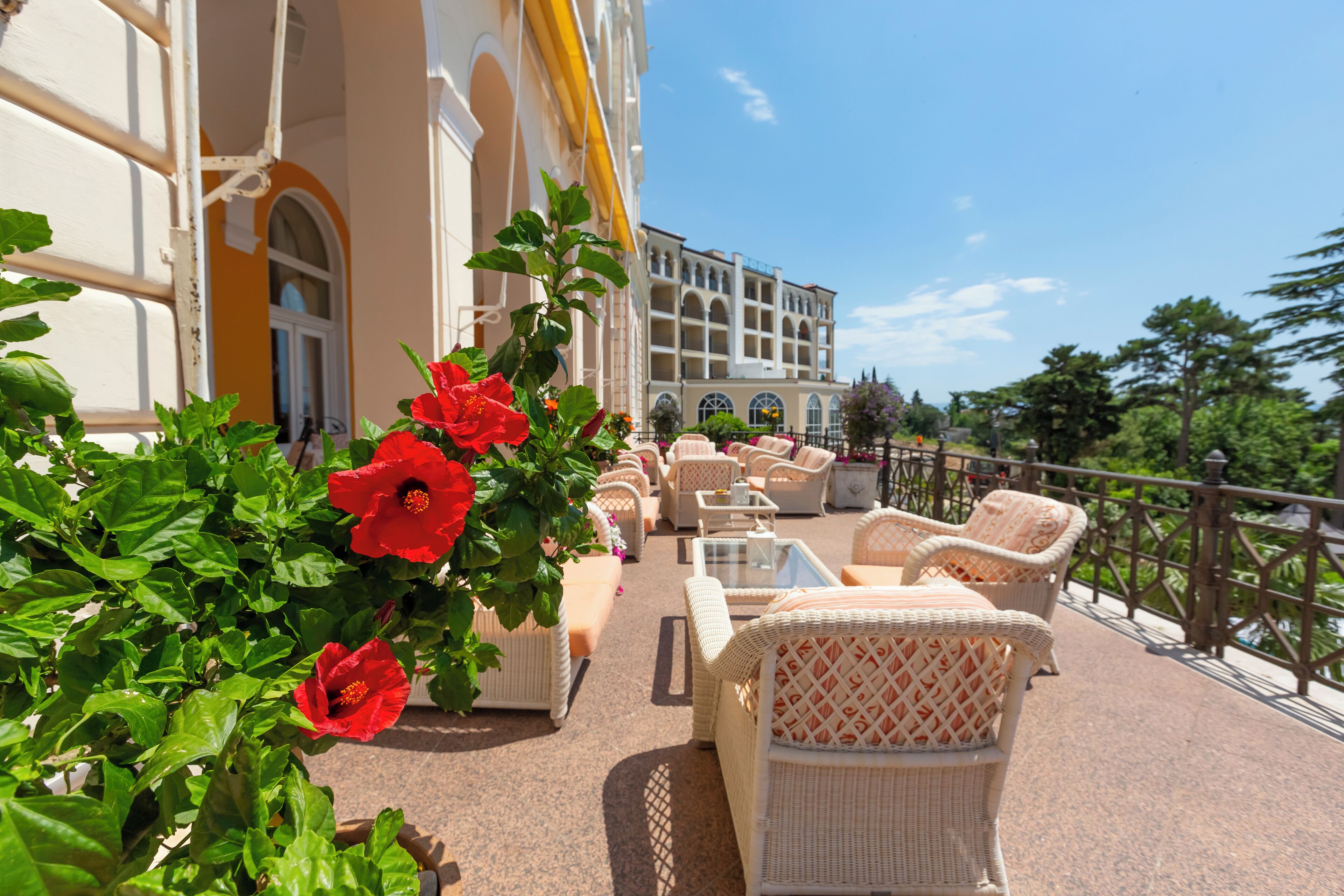 A sunlit outdoor seating area with wicker furniture and red flowers, overlooking a building with arched windows and a balcony.