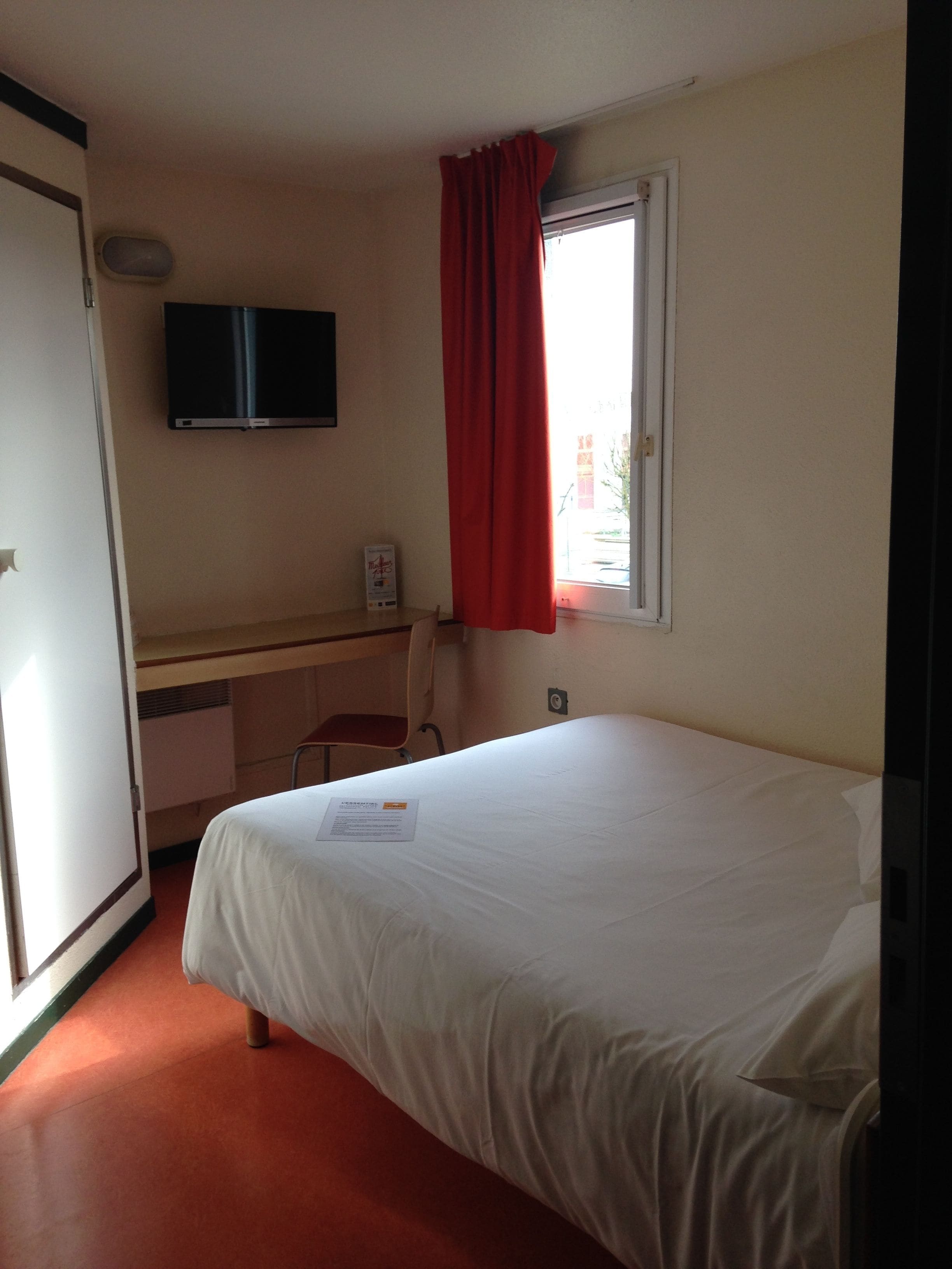 Ein Hotelzimmer mit Bett, Schreibtisch, Sessel, Fernseher und einem Fenster mit roten Vorhängen.