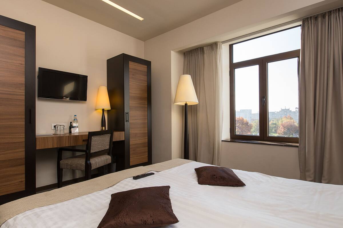Hotellihuone, jossa on sänky, kaksi tyynyä, työpöytä tuolilla, televisio ja ikkuna, jossa on verhot.