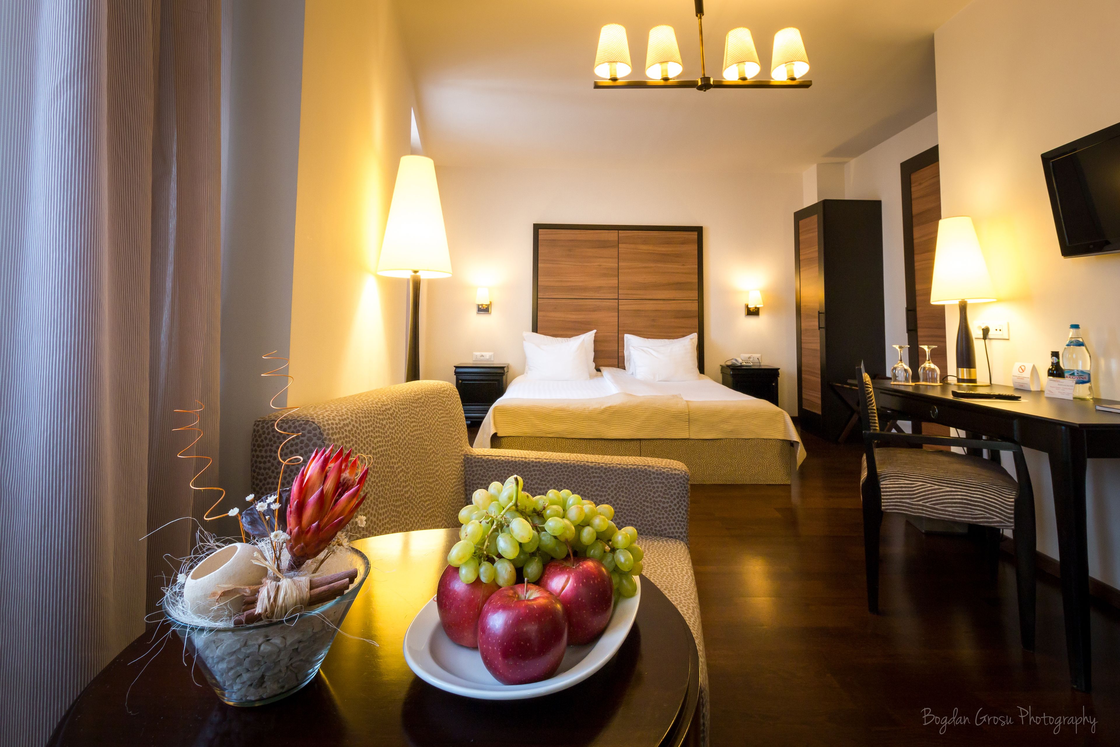 A hotel room with a bed, bedside tables, a desk, and a dining table with a fruit bowl.