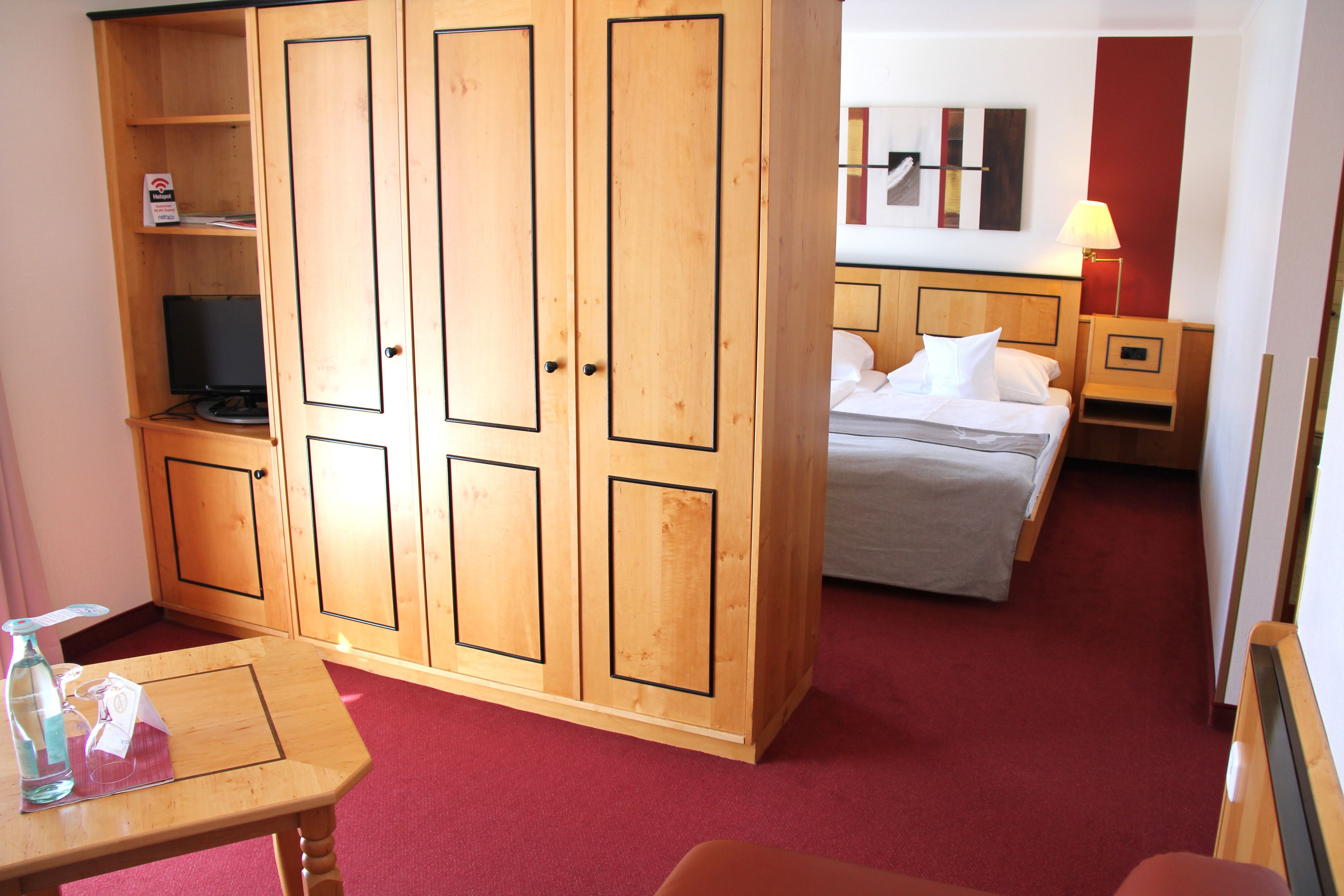 Una habitación de hotel con un armario de madera, una cama con ropa de cama blanca, una mesita con una botella y un vaso, y una alfombra roja.