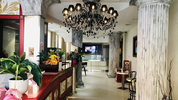 Un vestíbulo de hotel con mostrador de recepción, candelabro, columnas y plantas en macetas.