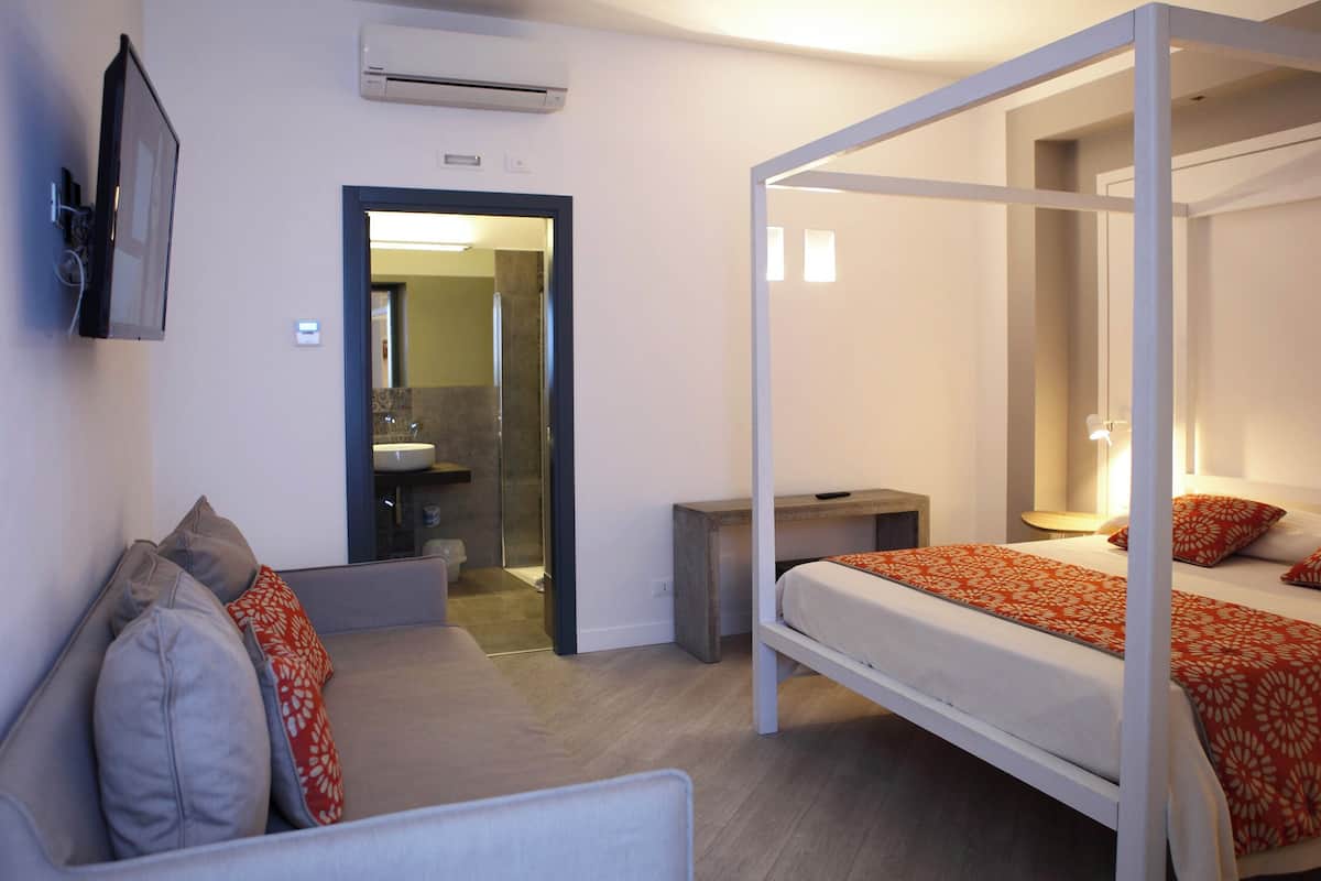 Habitación de hotel con sofá, cama con edredón estampado en naranja, mesita de noche y baño visible a través de una puerta abierta.