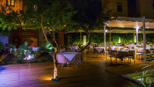 An outdoor dining area with tables and chairs, surrounded by trees and a building in the background.