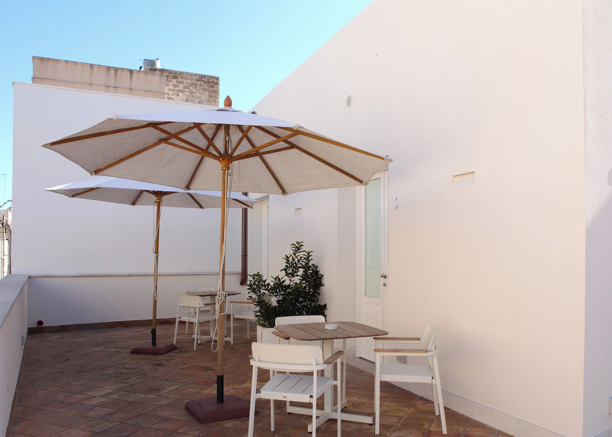 A small outdoor patio with two white tables and chairs, a large umbrella, and a potted plant.