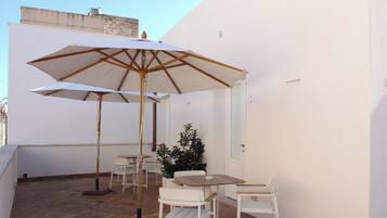 A small outdoor patio with two white tables and chairs, a large umbrella, and a potted plant.