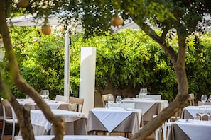 An outdoor dining area with tables set for two, surrounded by lush greenery and a white column. - Aegusa (Favignana)