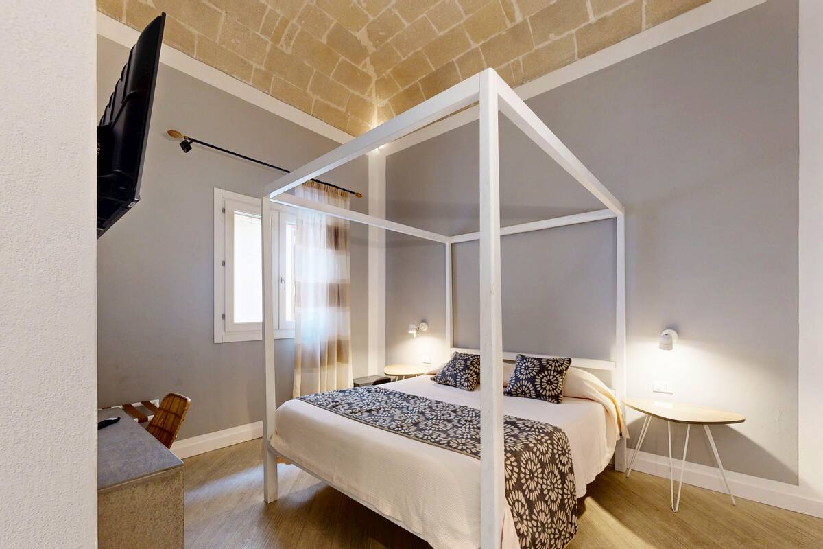 Un dormitorio con cama con dosel, una mesita de noche pequeña, una silla y una ventana con cortinas.