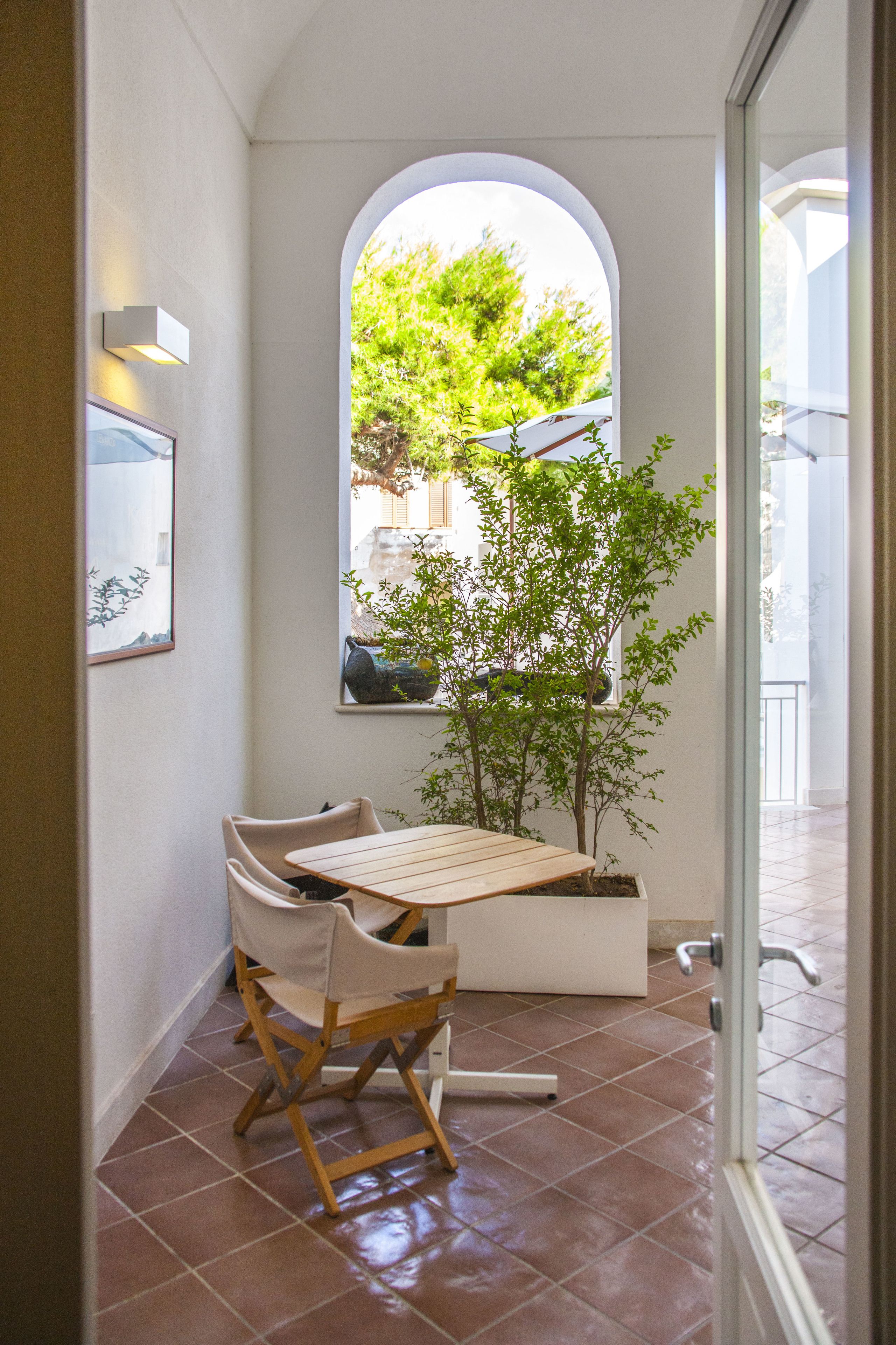 Un pequeño patio con una mesa, sillas, una planta en maceta y vistas a través de un ventanal arqueado.