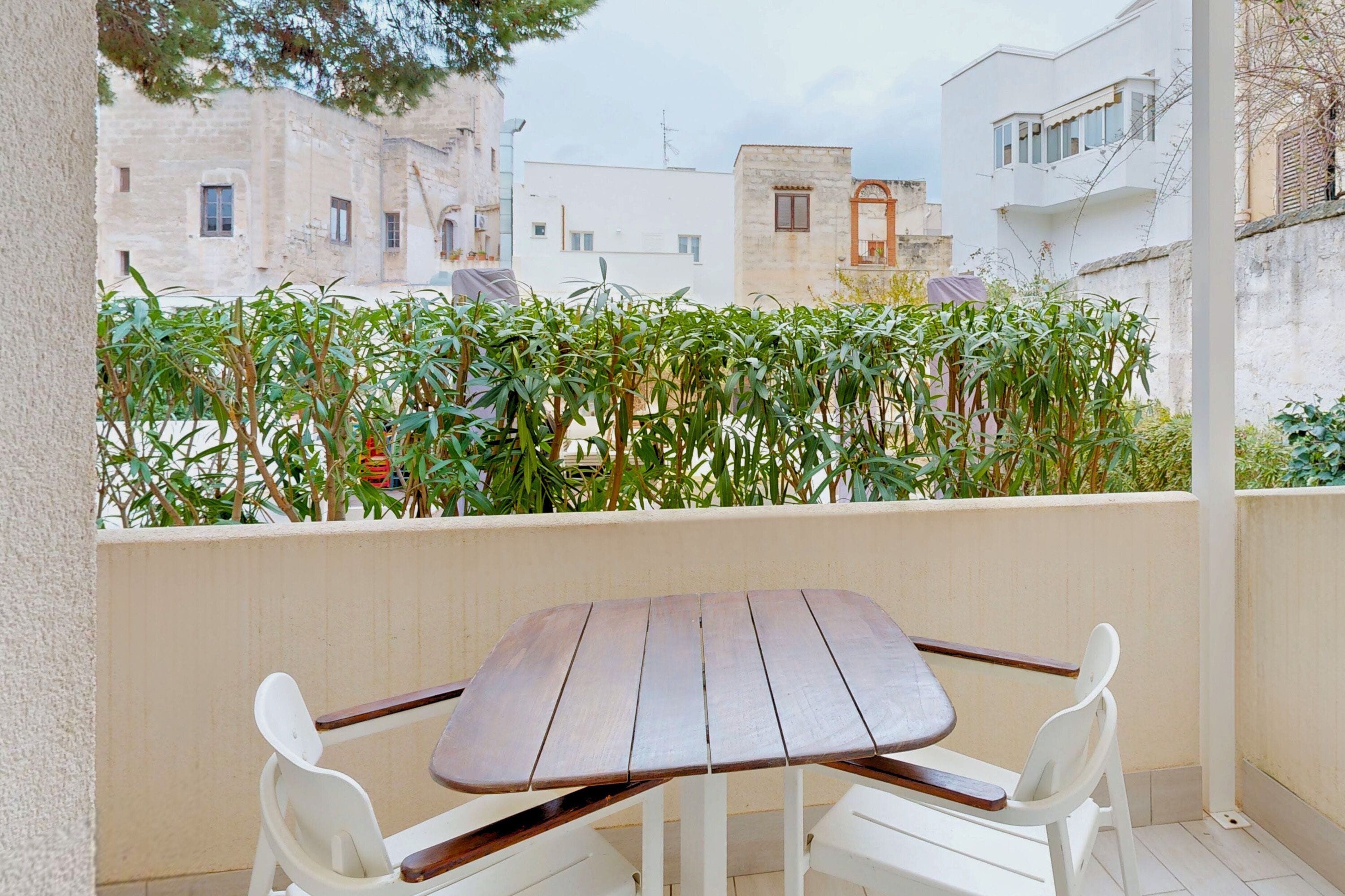 Un balcon avec une table en bois et des chaises blanches, donnant sur un quartier résidentiel avec des immeubles et de la verdure.