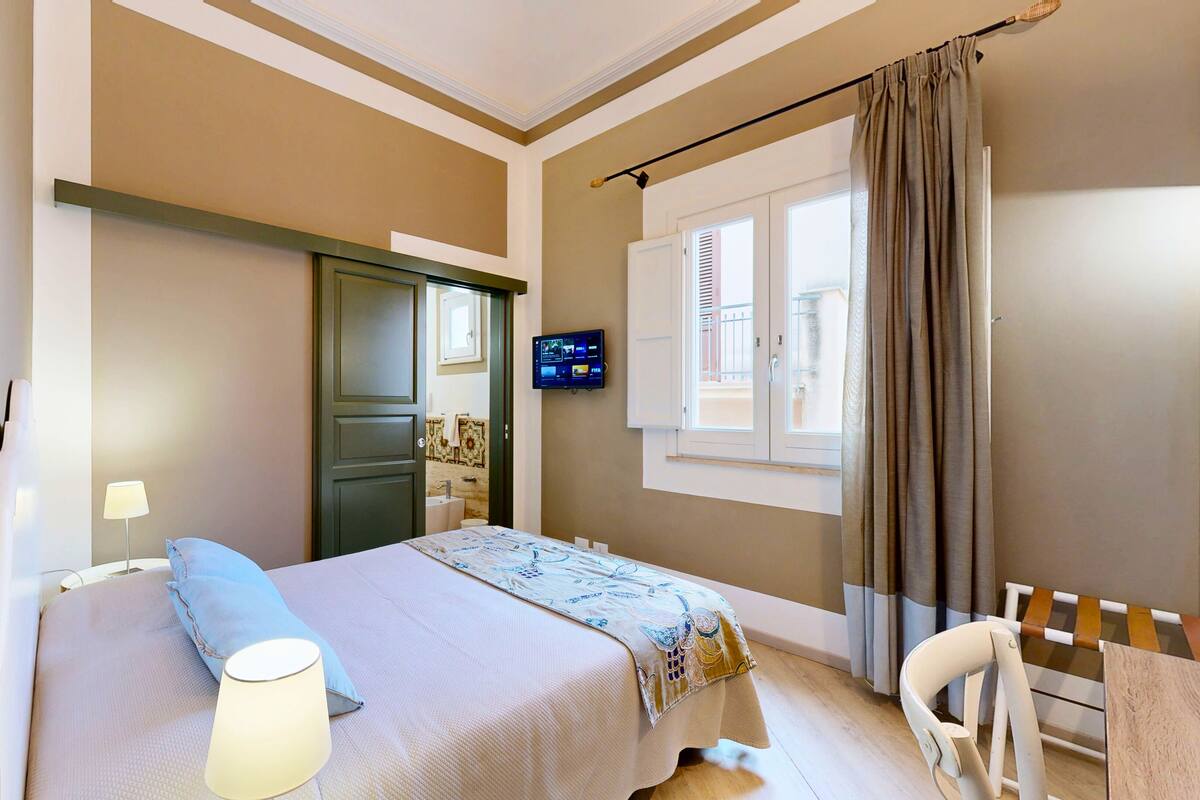 Un dormitorio con cama, televisor en la pared, ventana con cortinas y baño a la vista a través de una puerta abierta.