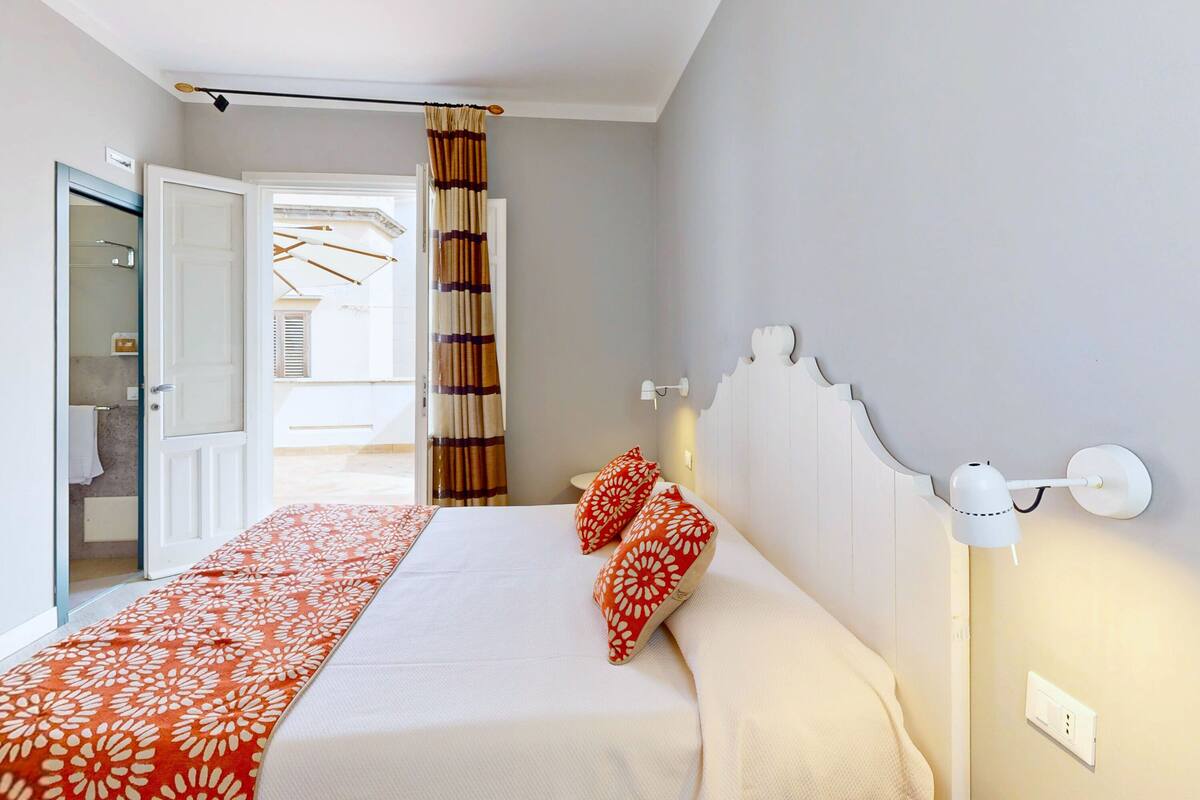 Un dormitorio con cama, dos almohadas, una mesita de noche, una lámpara de pared y vistas a un pasillo.