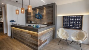 A modern reception area with a wooden reception desk, pendant lighting, and a wall-mounted display of jewelry. - Hotel Albert Plage (Knokke-Heist)