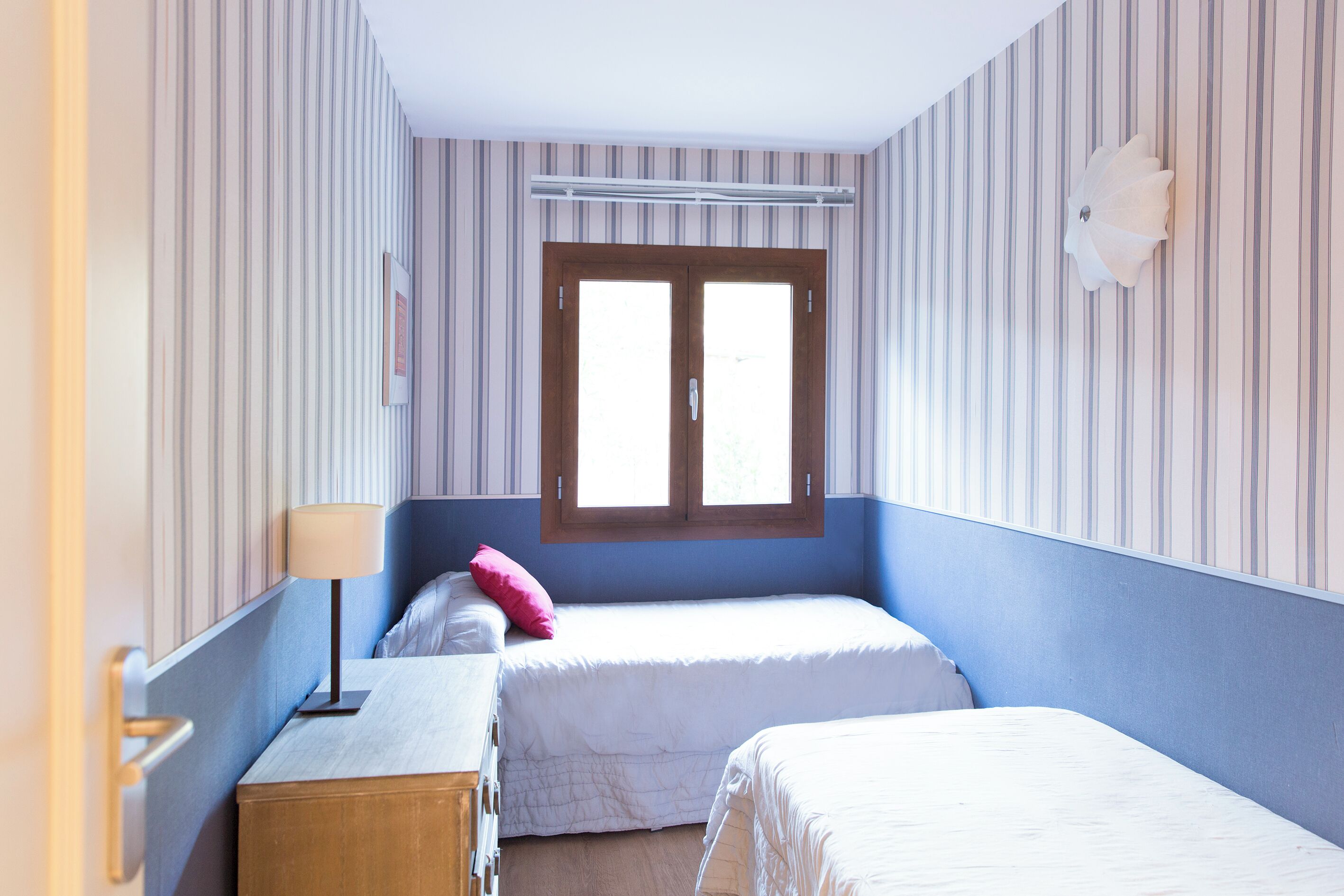 A room with two beds, a wooden dresser, a lamp, and a window with blinds.