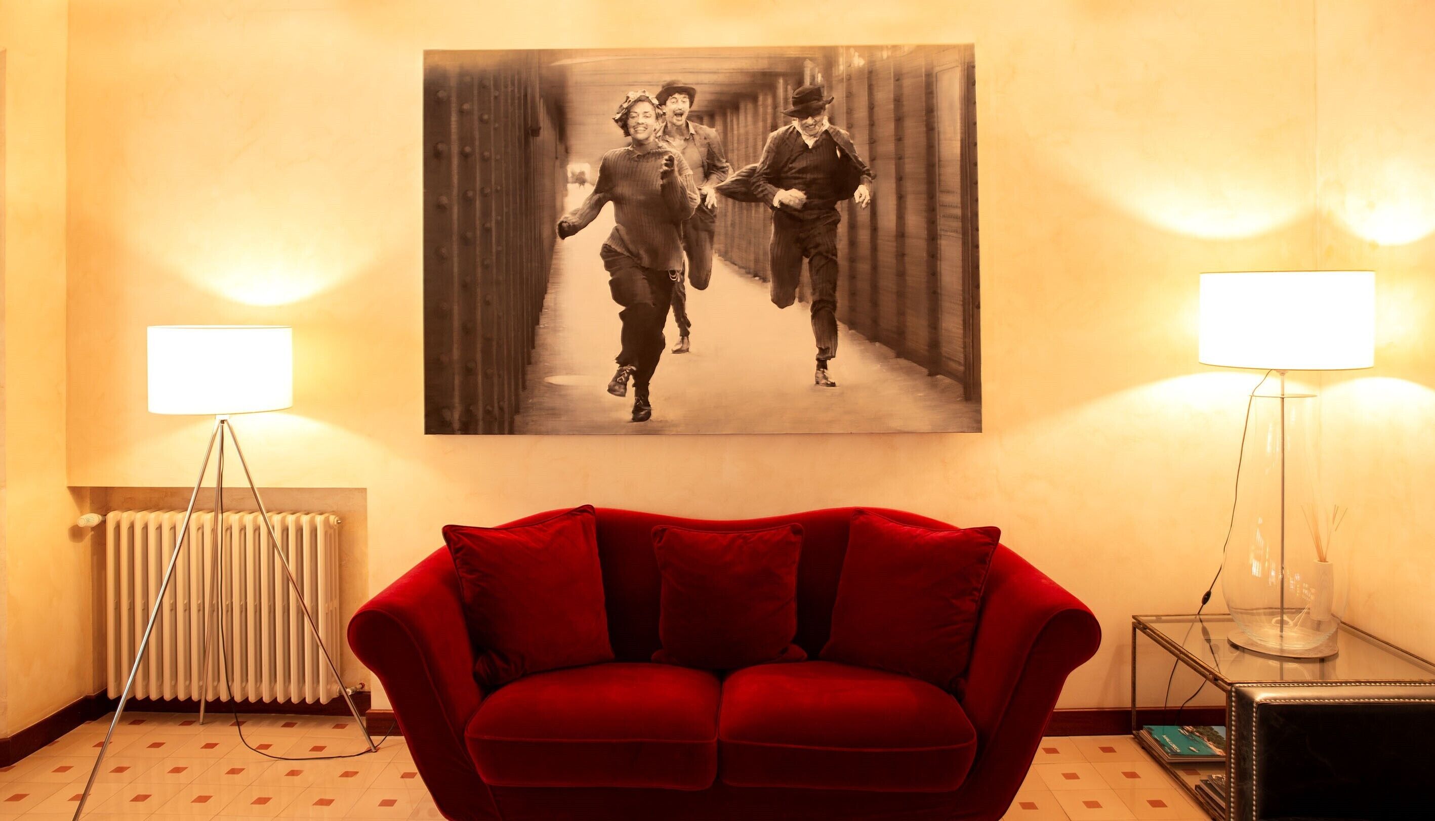 A room with a red sofa, a radiator, a floor lamp, and a painting of three people running.