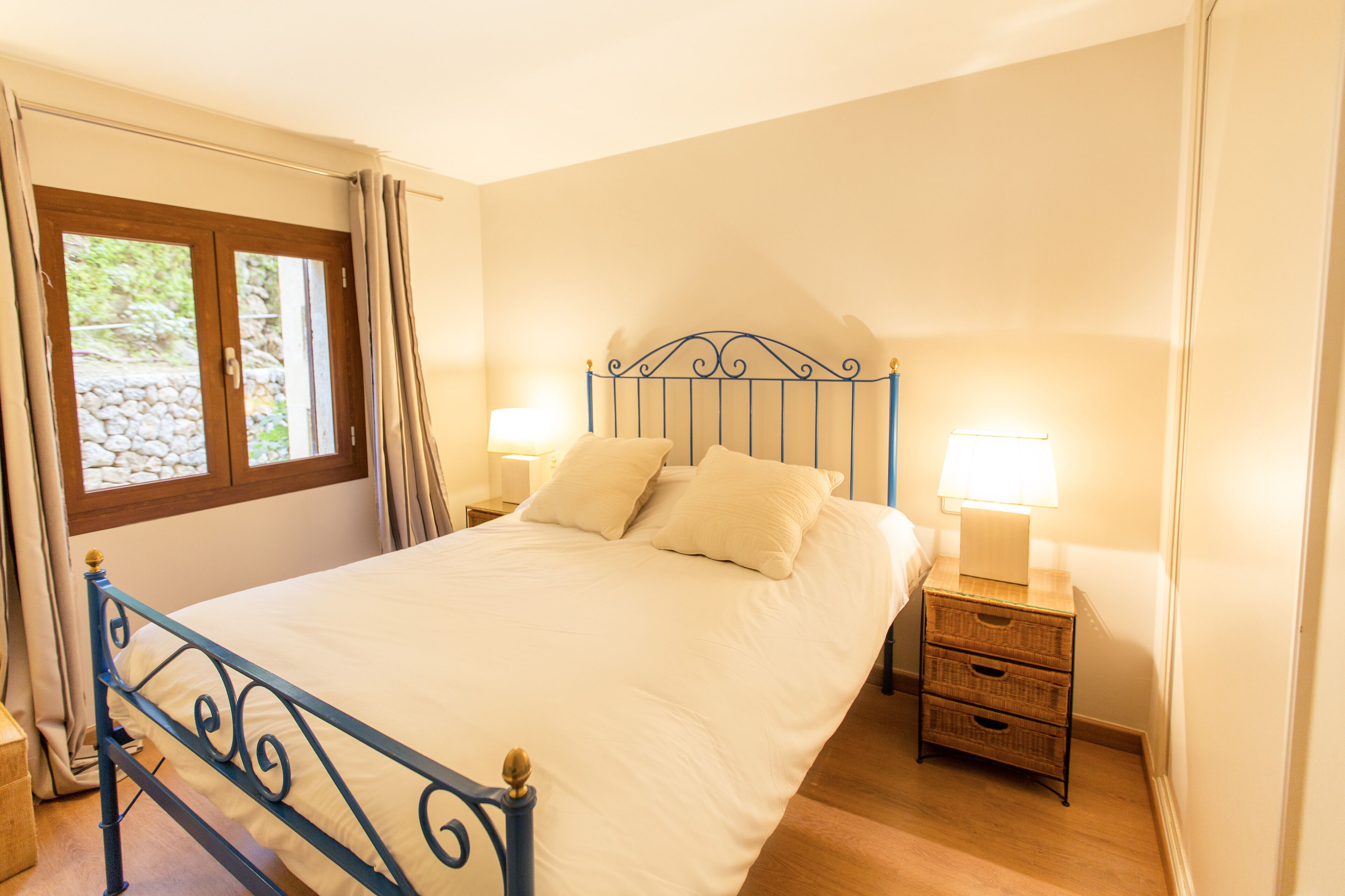 A bedroom with a bed, two bedside tables with lamps, a window with curtains, and a wooden nightstand.