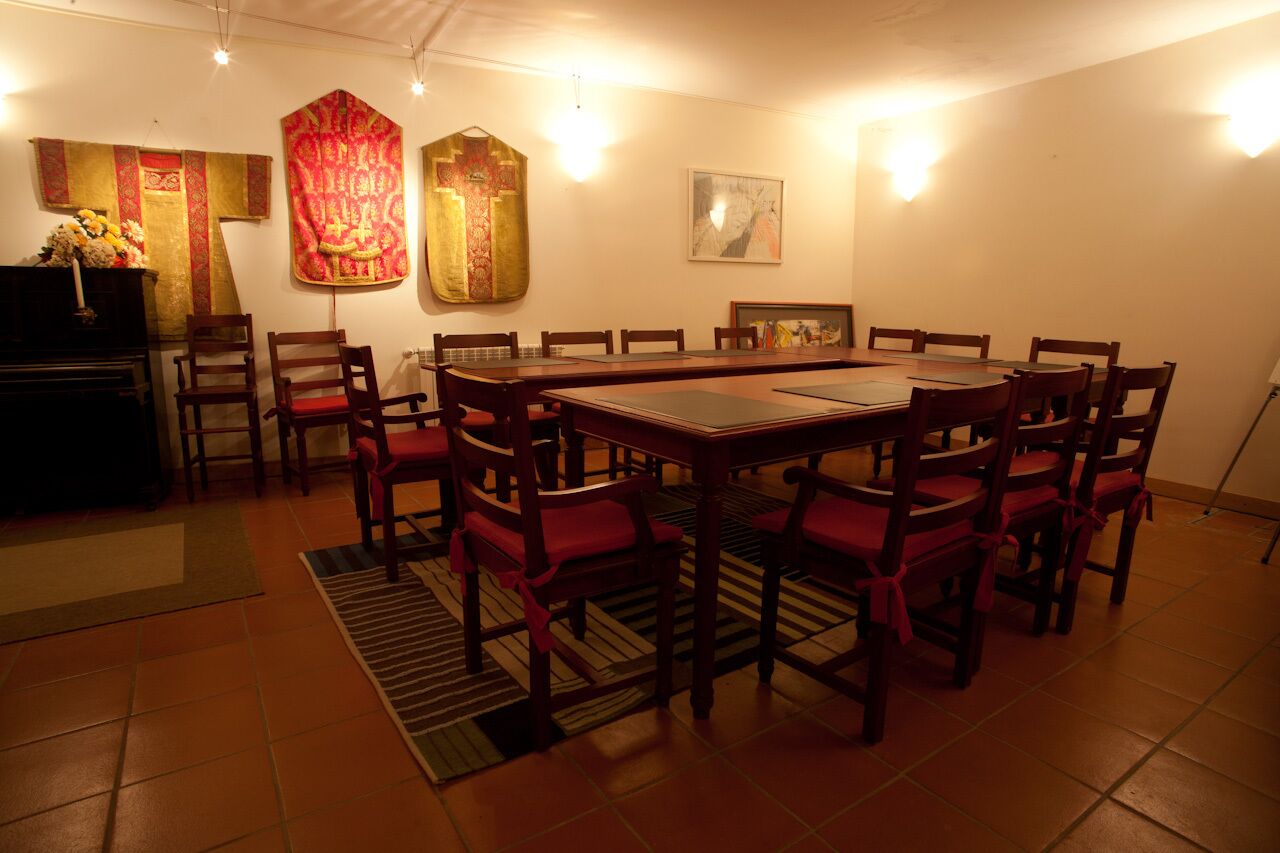 Un comedor con una amplia mesa rectangular rodeada de sillas de madera con cojines rojos. Se observan dos imágenes religiosas colgando en la pared, además de un cuadro enmarcado.