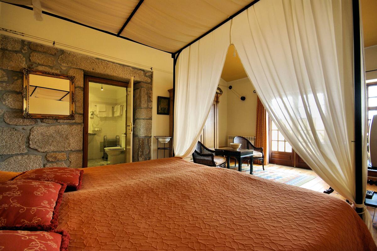 A bedroom with a stone wall, a bed with red pillows, a wooden headboard, and a large window with white curtains.