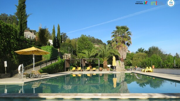 A clear swimming pool with lounge chairs and umbrellas, surrounded by greenery and a stone building in the background. - Hotel Rural Quinta da Geia (Oliveira do Hospital)