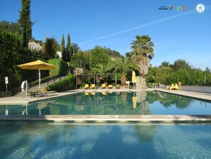 A clear swimming pool with lounge chairs and umbrellas, surrounded by greenery and a stone building in the background.