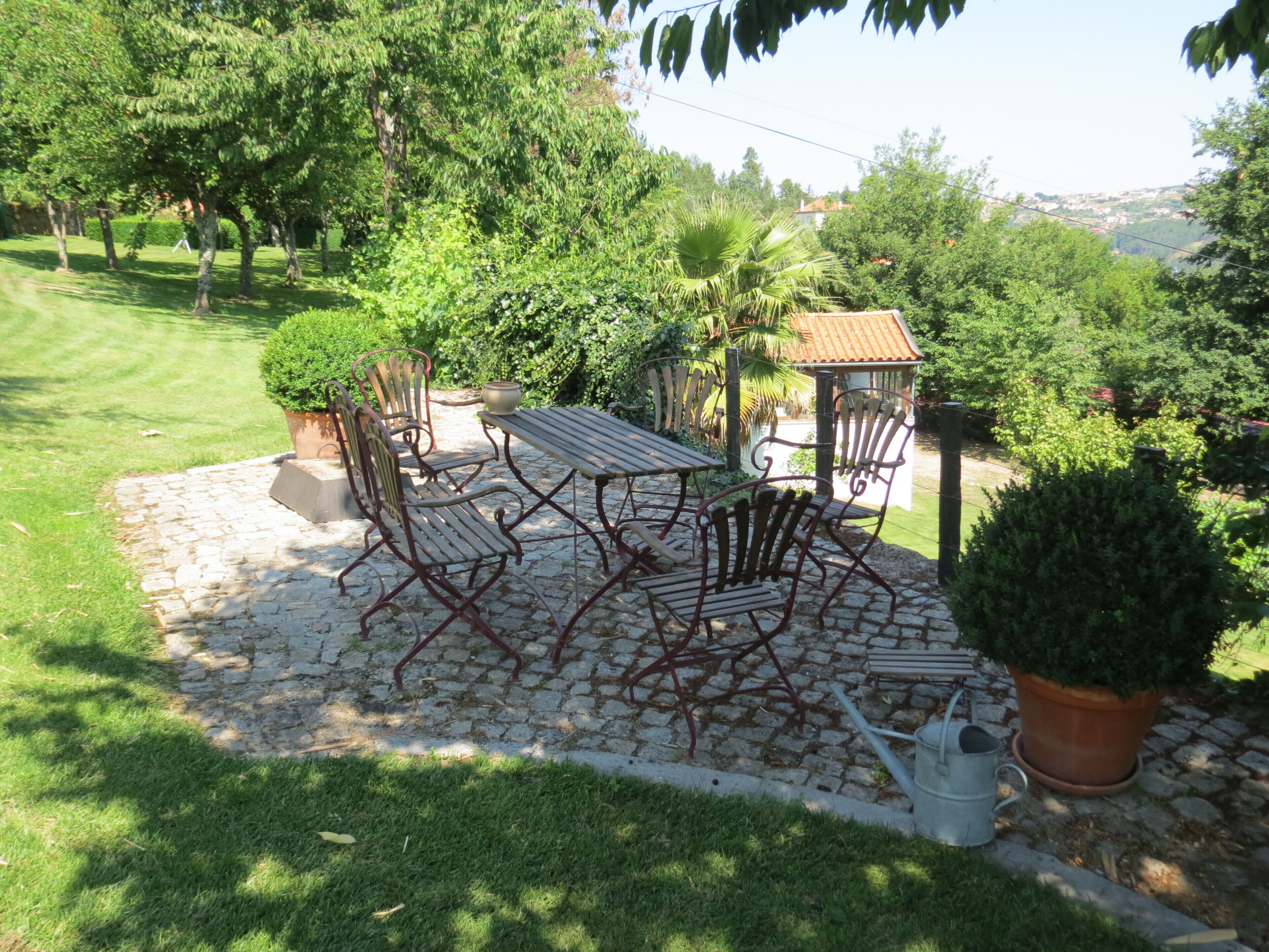 A patio with a table and chairs, surrounded by greenery and a cobblestone path.