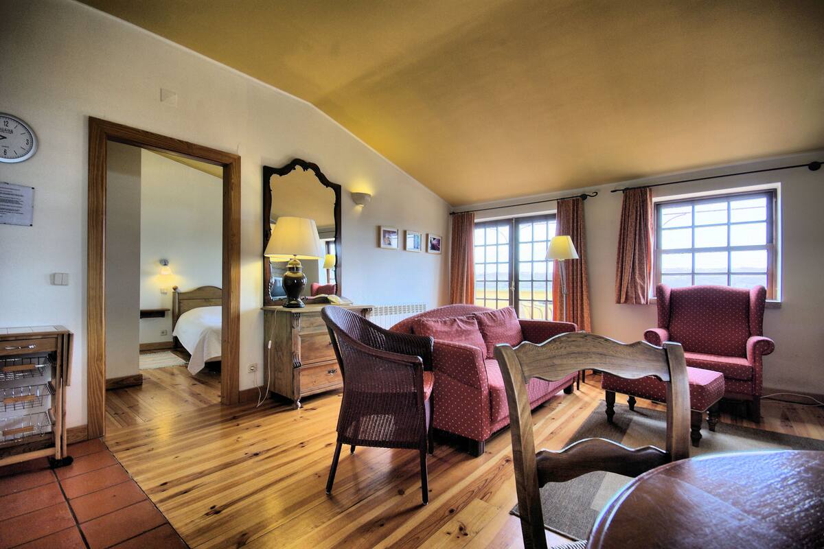 A room with a wooden floor, a red sofa, a wooden chair, a wooden bed, a mirror, and a window with curtains.