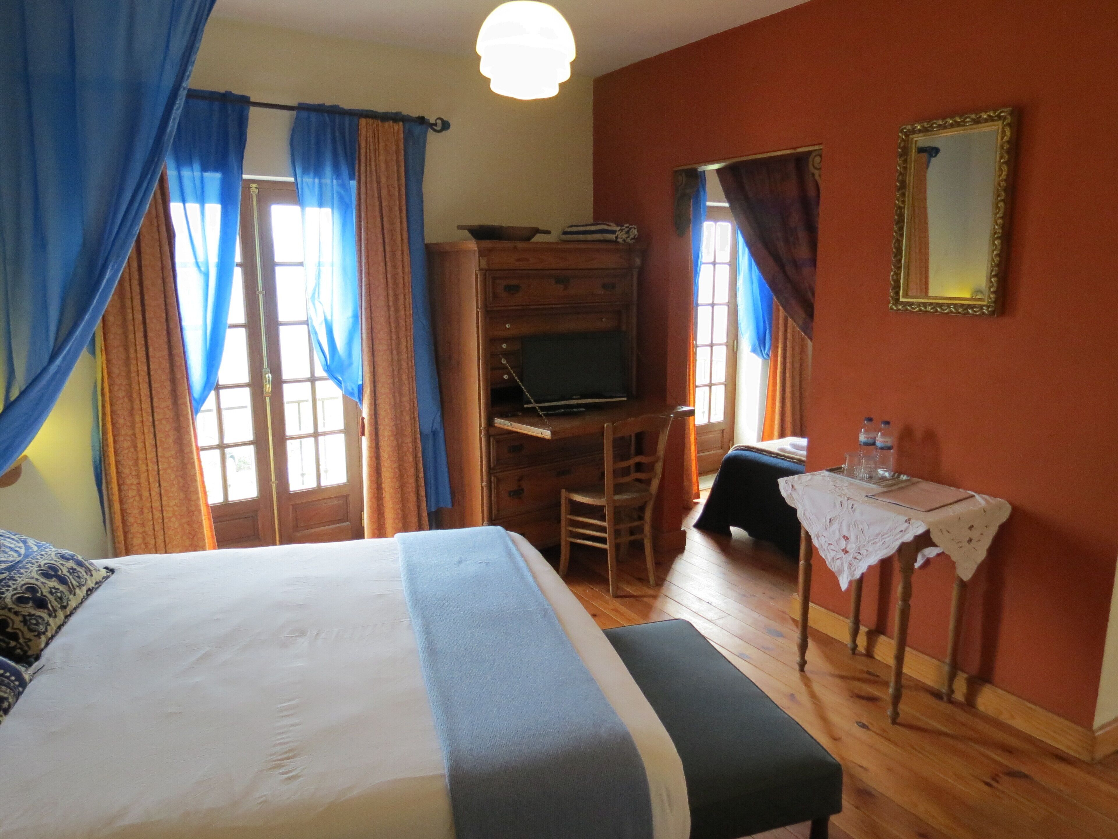 A bedroom with a bed, a desk, a chair, a small table with a lamp, and curtains.