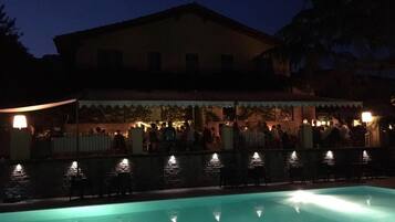 Ein Poolbereich mit einem Gebäude im Hintergrund, einer überdachten Sitzgelegenheit und einem gut beleuchteten Pool.