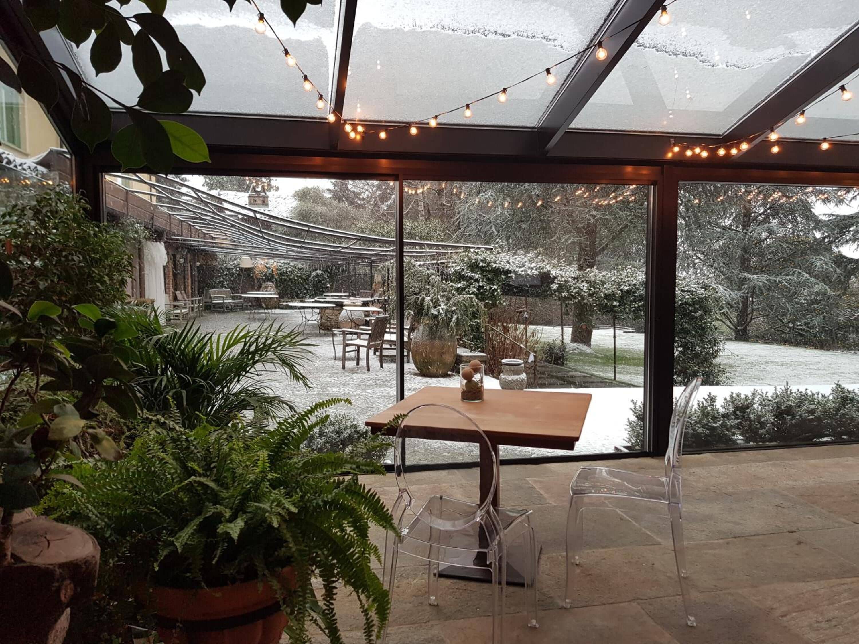 Ein verglaster Patio-Bereich mit Tisch und Stühlen, umgeben von Topfpflanzen und Blick auf einen verschneiten Garten.