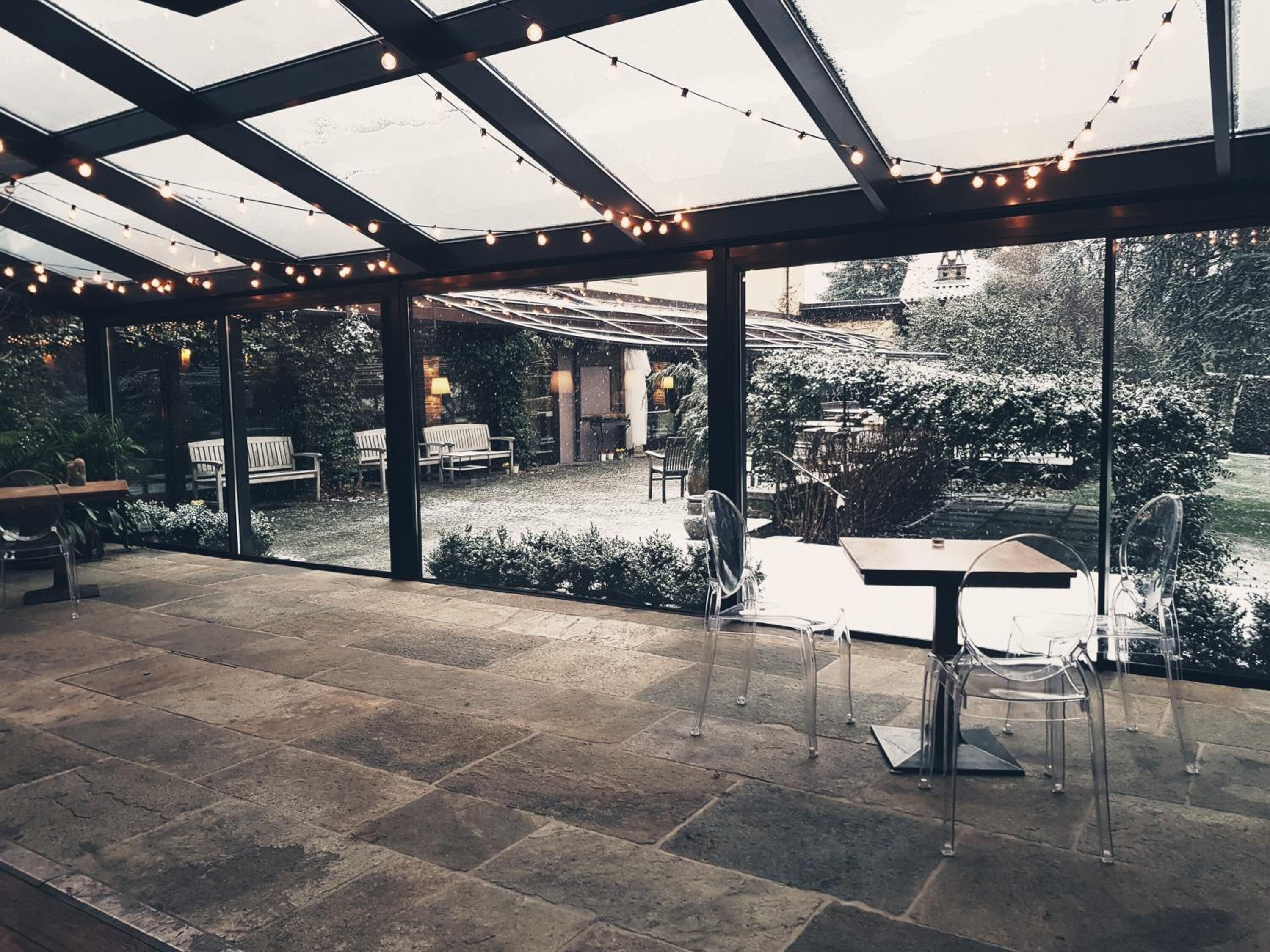A covered outdoor patio with stone flooring, clear glass walls, and string lights.