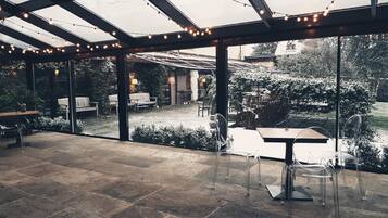 A covered outdoor patio with stone flooring, clear glass walls, and string lights.
