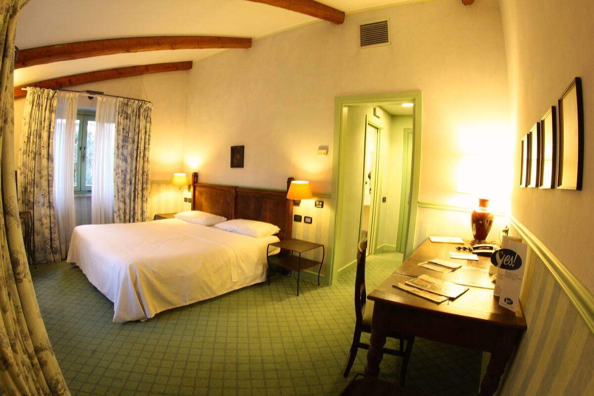 A hotel room with a bed, a desk, a chair, a lamp, and a door.