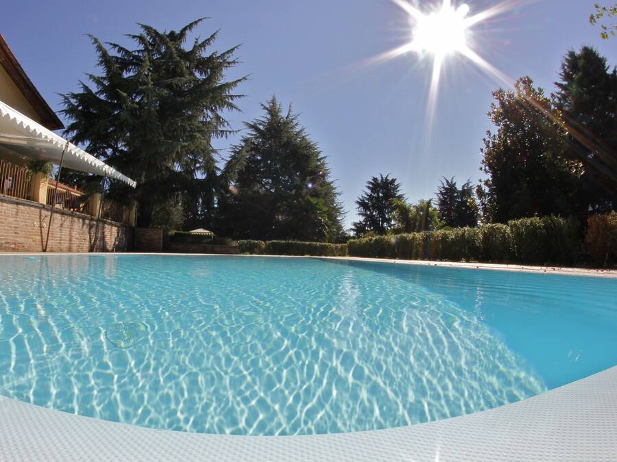 Una piscina cristalina con un borde de baldosas, rodeada por un muro de ladrillo, árboles y un edificio con toldo blanco.
