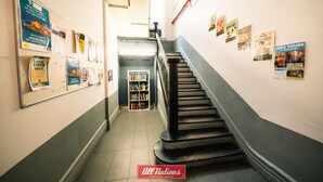 Un pasillo con una escalera, carteles en las paredes y una estantería al fondo.