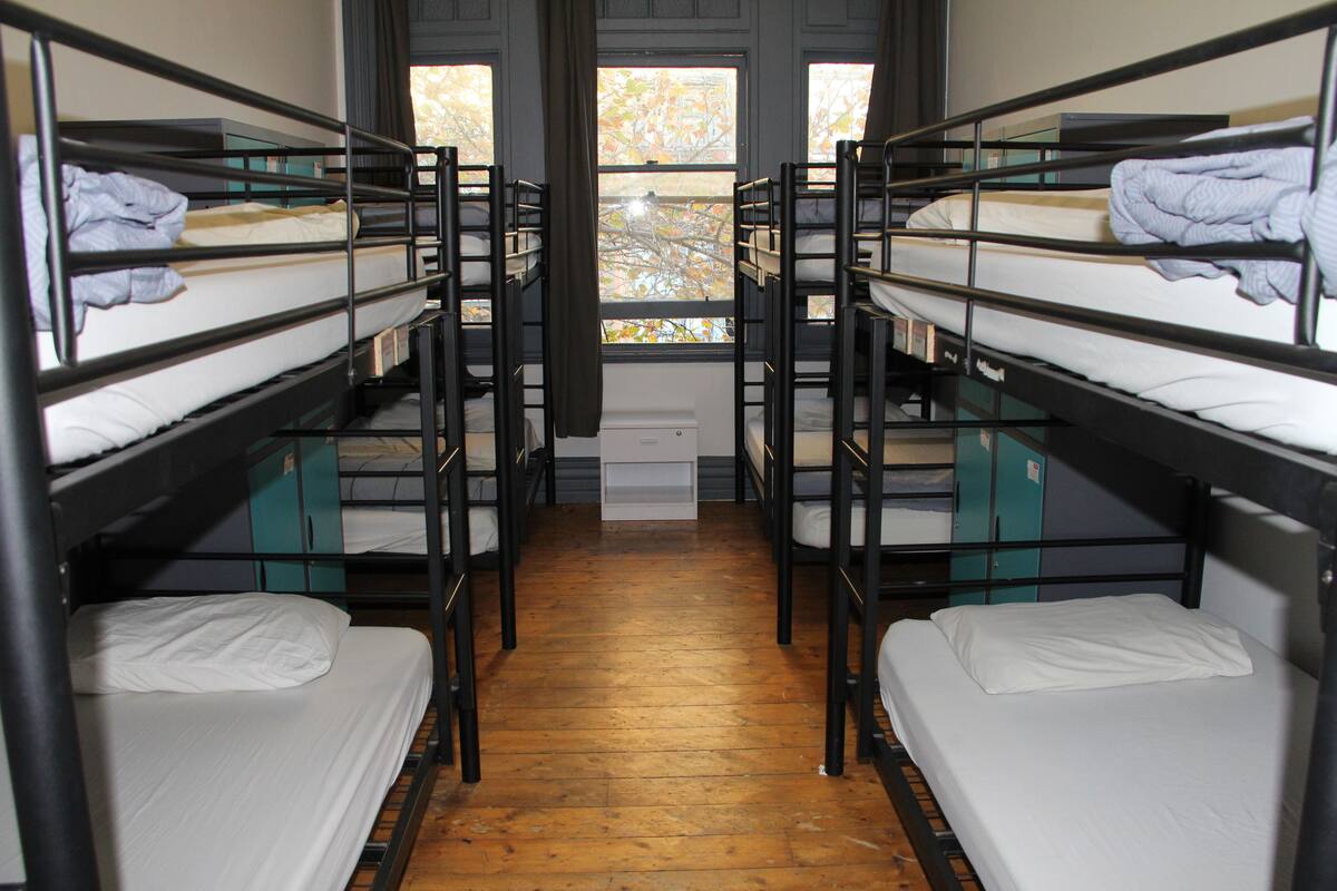 A dormitory room with bunk beds, wooden flooring, and a window with a view of trees.