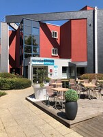 Um edifício moderno com fachada vermelha, entrada de vidro e área externa com cadeiras e mesas de vime.