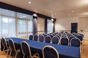 Uma sala de conferências com uma mesa comprida e fileiras de cadeiras, grandes janelas com cortinas, e uma porta com placa de saída de emergência.