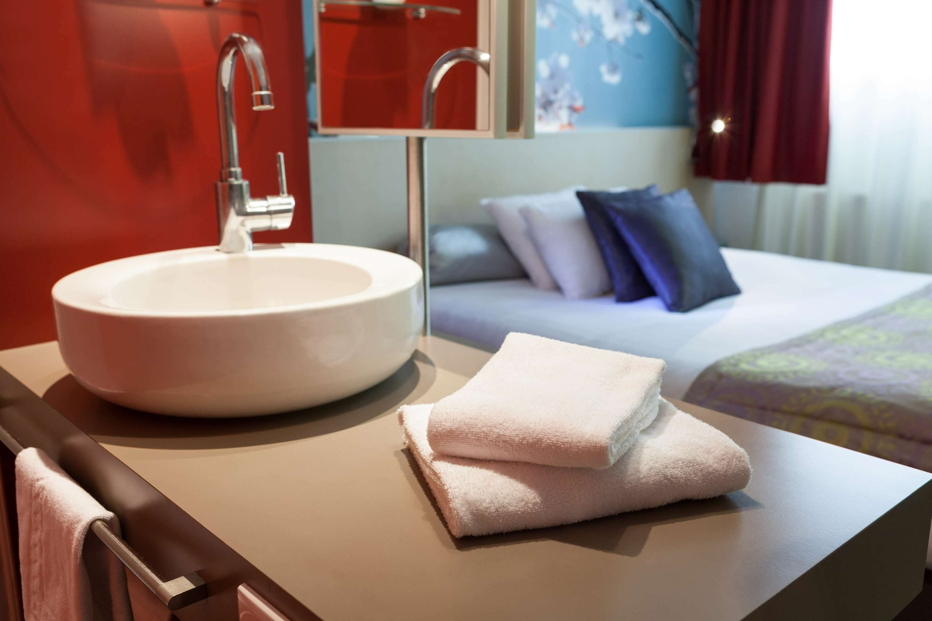A hotel bathroom with a round sink, a towel, and a bed with pillows.