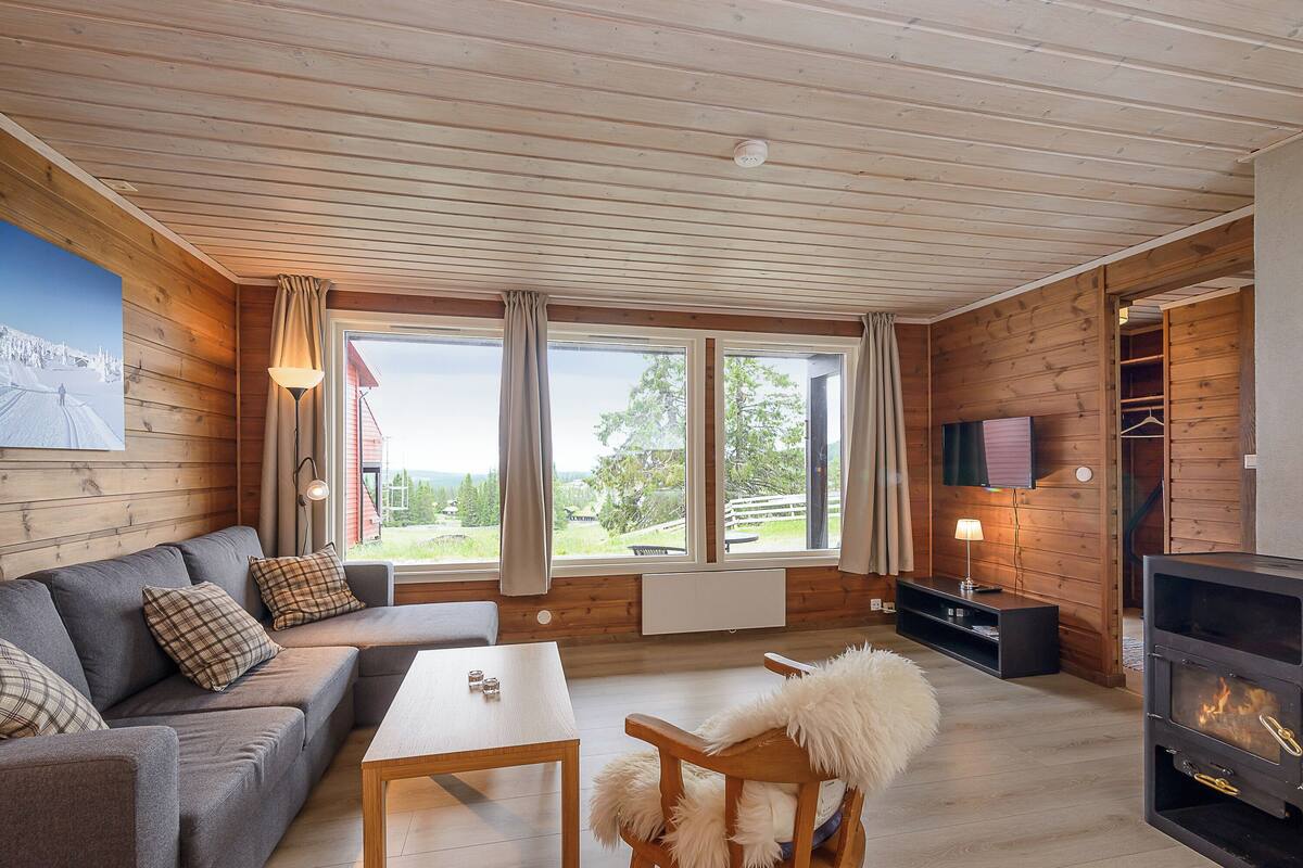 Ein gemütliches Zimmer mit Holzbalkendecke, einer Couch, einem Tisch, einem Stuhl, einem Kamin und großen Fenstern.