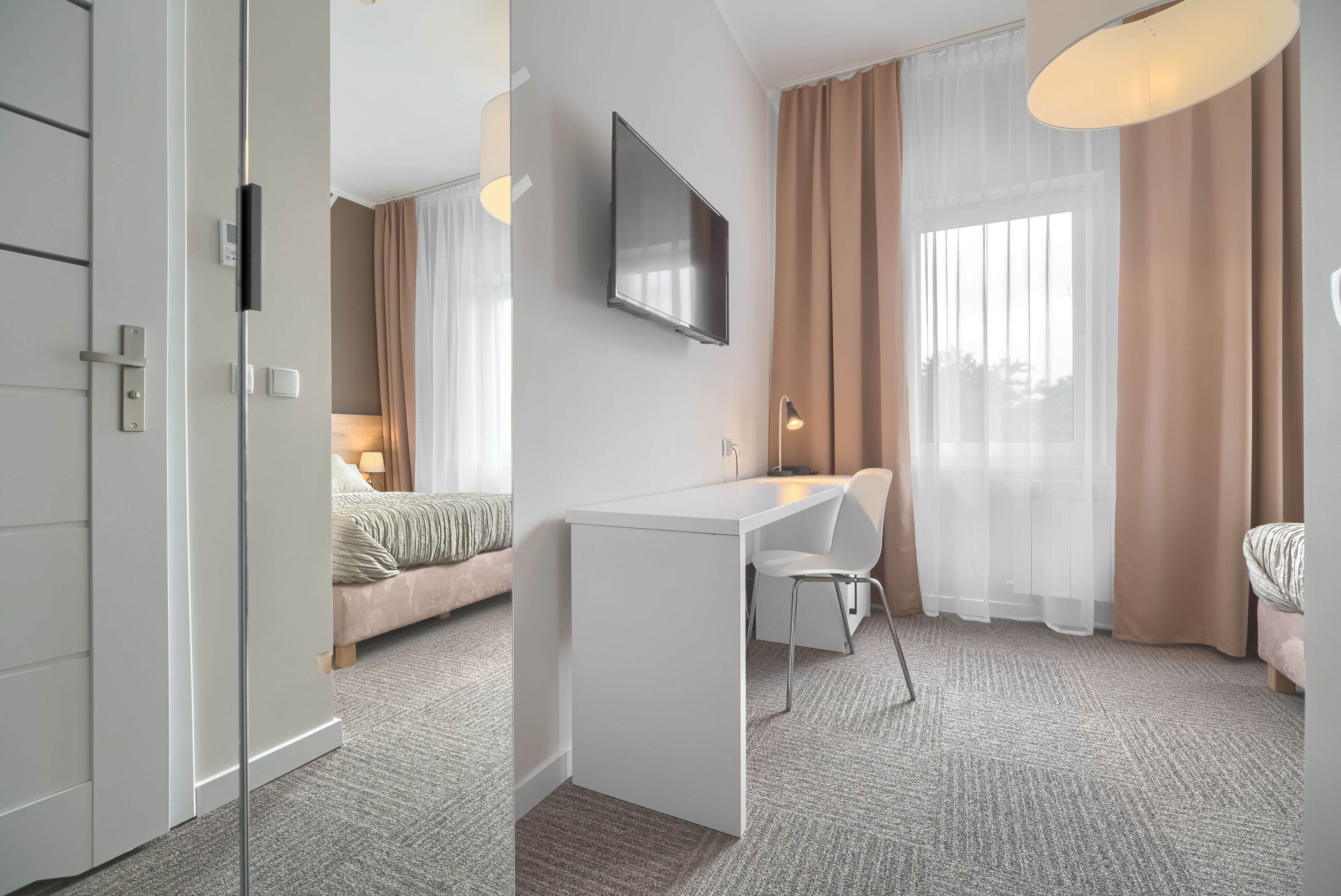 A modern hotel room with a white desk, a flat-screen TV, a bed with a beige headboard, and a large window with sheer curtains.