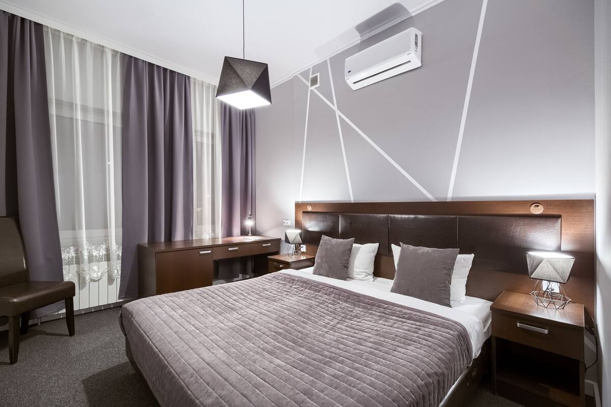 A modern hotel room with a large bed, a desk, and a chair. There is a window with curtains, a wall-mounted air conditioner, and a pendant light above the bed.