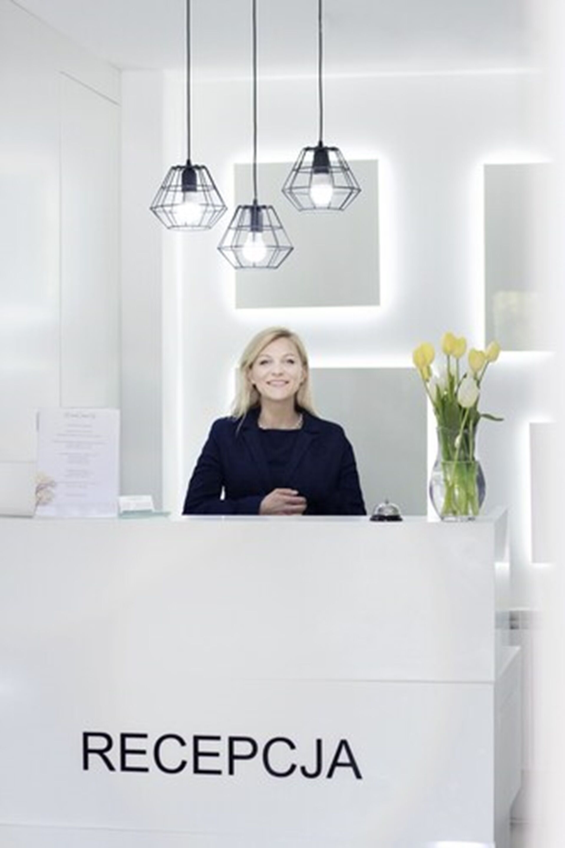 A reception area with a white counter labeled "RECEPTCJA," a receptionist, and a vase with yellow flowers.